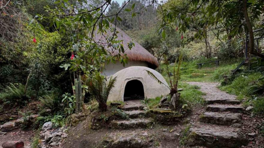 Primer plano de Maloca rodeada de árboles y vegetación. Hay una especie de gruta al frente y camino piedra alrededor.