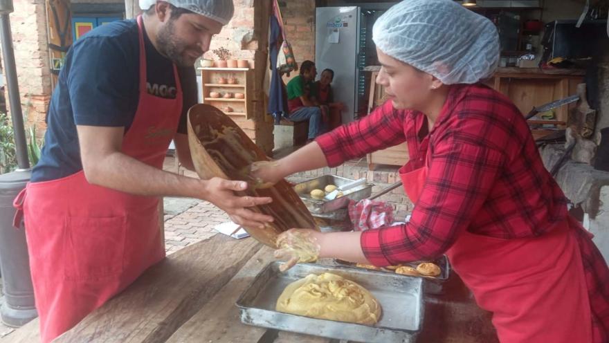  Un hombre y una mujer jóvenes cocinan con delantal y cofias unos amasijos. La cocina es artesanal.