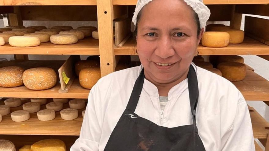 Mujer joven de tez canela sonriente con cofia delante de una estantería de madera con quesos.