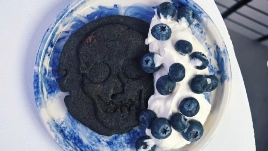 Postre con galleta, helado y arándanos. La galleta es negra y tieje forma de calabera. El postre está sobre un plato blanco con manchas azules.