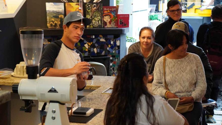 Grupo de personas jóvenes observan a un joven que prepara café. hay una máquina de preparar café sobre una mesa.