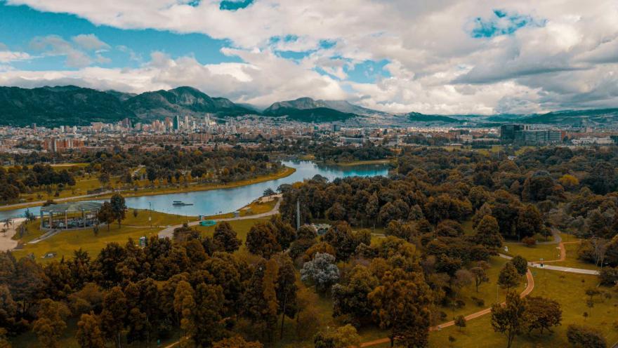 Parque Simón Bolívar plano general desde arriba en dron, se ven las coronas de los árboles, amplias zonas verdes, el gigantesco lago, edificios de la ciudad al fondo y los cerros orientales bajo un cielo azul nubado.