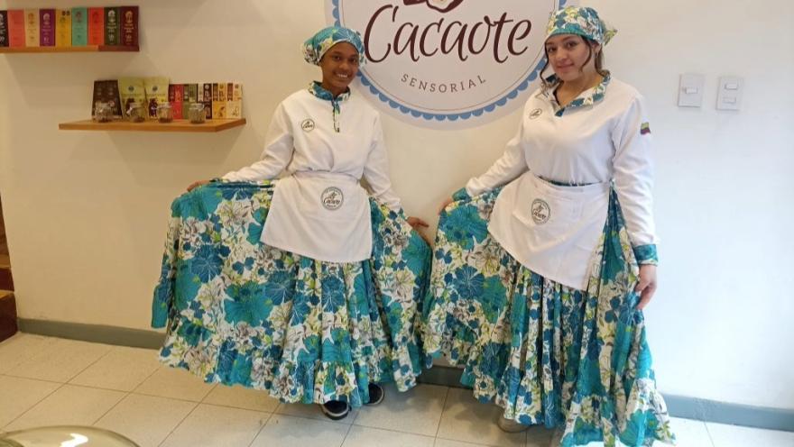 Dos mujeres jóvenes vestidas con delantales de color azul y blanco donde resaltan varias leyendas que dicen "Cacaote"