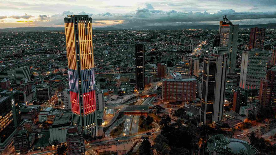 En el atradecer iluminada la Torre Colpatria con los colores de la bandera de Colombia. Al fondo las luces de la inmensa ciudad en la que destacan algunos edificios altos y el Planetario.