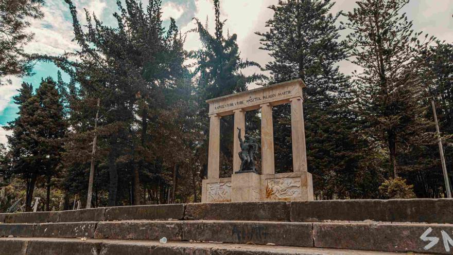 Monumento en piedra tallada con una estatua negra al frente y varios pinos altos detrás.