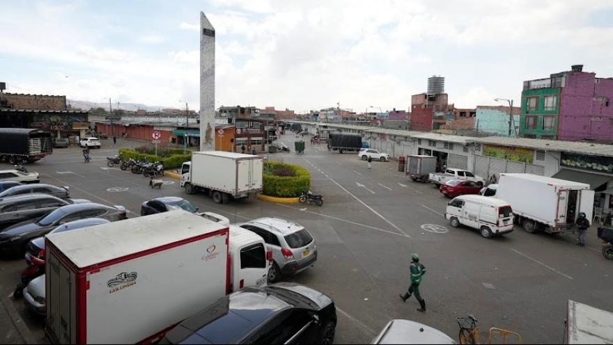 Corabastos, plano general de una gran plazoleta, rodeada de locales comerciales. Hay varios caminones alrededor. Hay un obelisco blanco en la mitad.