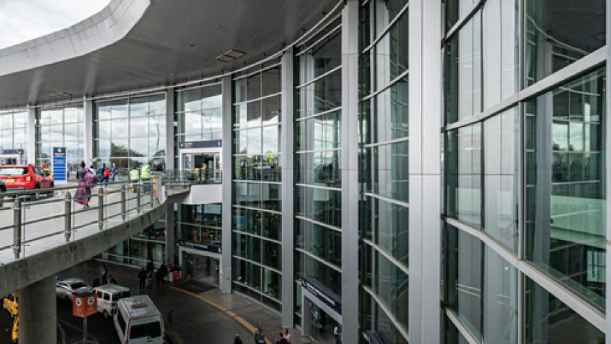 Exterior of a two-story building. Several vans are parked on the ground floor. On the second floor, there is a pedestrian bridge with some people walking across it.