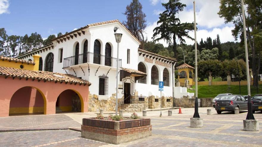 Museo Arqueológico fachada estilo colonial de coor blanco. Al frente una plazoleta con algunos vehículos estacionados y al fondo árboles altos.