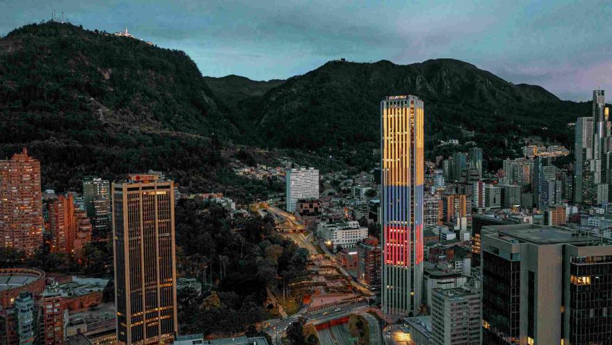 En el atardecer iluminada la Torre Colpatria con los colores de la bandera de Colombia y vista de occidente a oriente con algunos edificios altos y los cerros orientales de fondo.