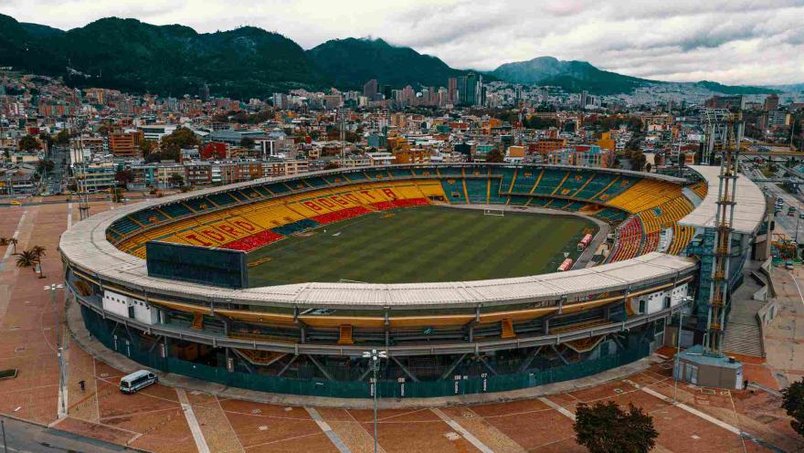 Estadio El Campín en primer plano visto de norte a sur con la ciudad de fondo. El cielo está cubierto de nubes