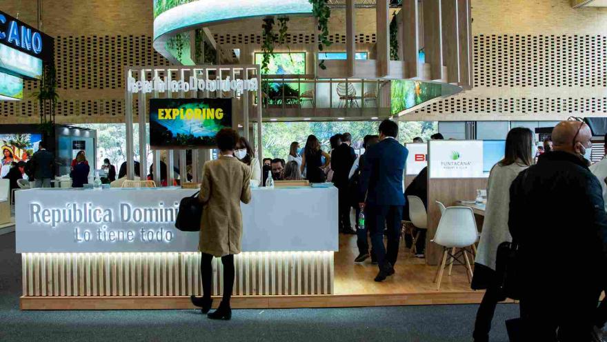 Salón de eventos con un gran stand en el que se lee "República Dominicana". Hay personas alrededor.