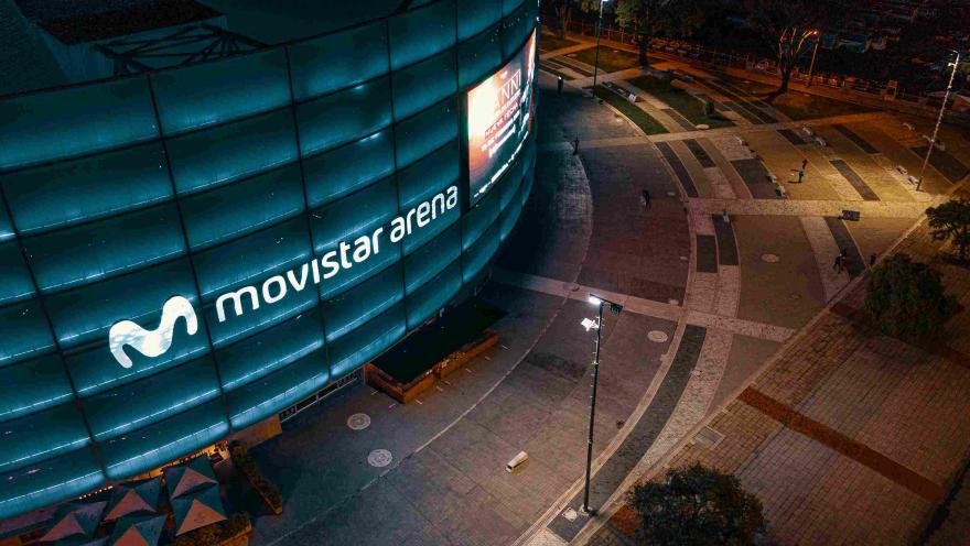 Fachada del movistar Arena de noce con escenario iluminado en azul y con el aviso con letras en neón color blanco. Al frente hay una plazoleta en ladrillo, concreto y vegetación.