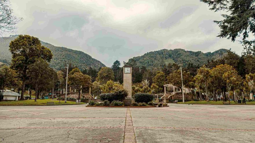 De día una amplia plazoleta con una torre que tiene un reloj en su parte alta. El lugar está rodeado de árboles y al fondo los cerros orientales.