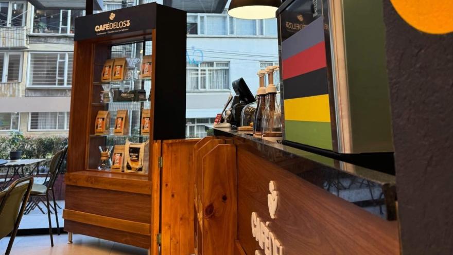  Interior de un local de día en el que se ve a un lado una barra con accesorios para hacer café, un anaquel de madera con café en bolsas. Hay unas mesas con sillas alrededor y al fondo un gran ventanal hacia la calle donde se ve la fachada de un edificio.