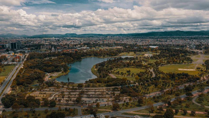 Parque Simón Bolívar plano general desde muy arriba en dron de oriente a occidente, se ven zonas verdes, árboles alrededor, el lago en el centro y al fondo edificaciones de la ciudad. El cielo está tapado de nubes.