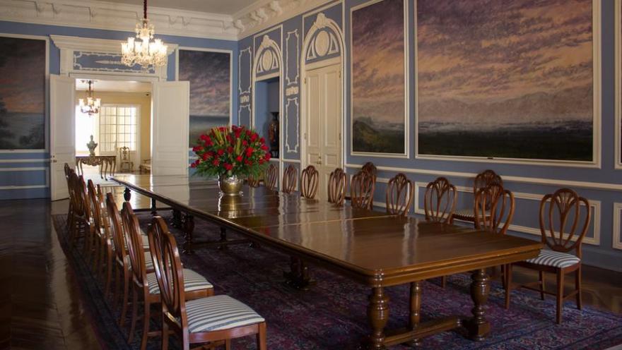 Salon pintado de azul en el que destaca una mesa larga de madera rodeada por sillas, también de madera. En la mesa hay un gran jarrón con flores rojas.