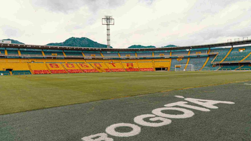 Interior del Estadio, desde el suelo, se observa una tribuna de fondo pintada con la palabra Bogotá. Hay una publicidad en el suelo también con la palabra Bogotá pintada sobre una gramilla artificial. Al fondo se alcanzan a ver los cerros orientales. 