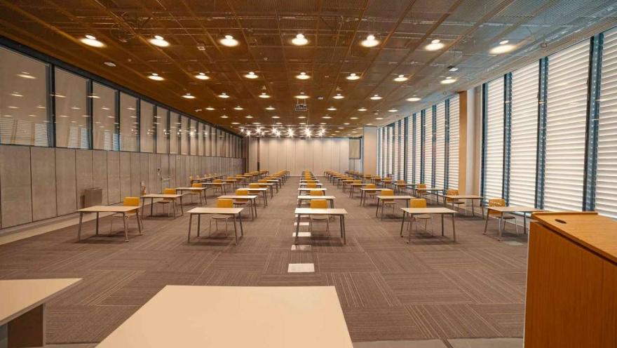 Salón de eventos con grandes ventanales a los lados, luces en el techo, piso entapetado y varias mesas con sillas alrededor.