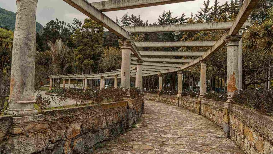 Estructura en piedra tallada y grandes columnas representando una pérgola dividida en dos semicírculos, y rodeada completamente de árboles.