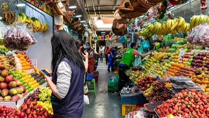 Pasilllo dentro de plaza de mercado con locales atiborrados de frutas. Hay varios vendedores al frente de cada local.