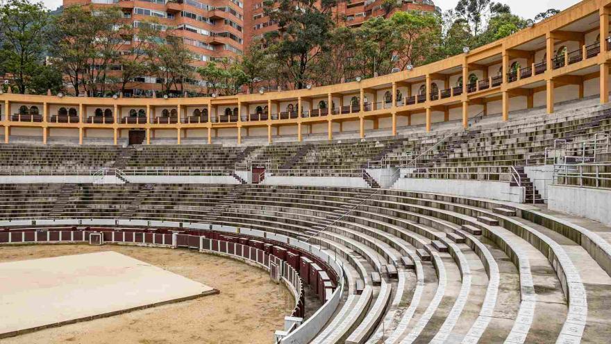 Interior de la plaza, se ve un costado de una gradería blanca ovalada de dos pisos y un tercero con balcones de color amarillo. Por encima y al fondo resaltan dos torres de edificios de ladrillo y unos árboles altos.