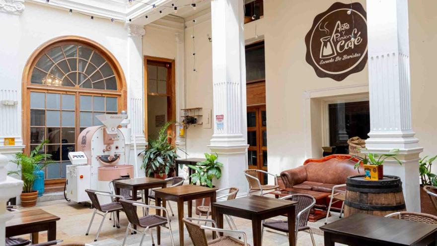 Salón principal de un café en el que se ven mesas de madera y sillas de mimbre, 3 columnas de mármol que sostienen el lugar y le dan un elegante decorado y un aviso en una pared que dice "Arte y pasión café".