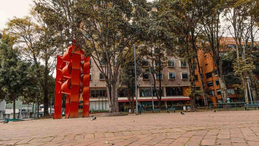 A tall, red metal sculpture in a small plaza with the facades of several buildings visible in the background.