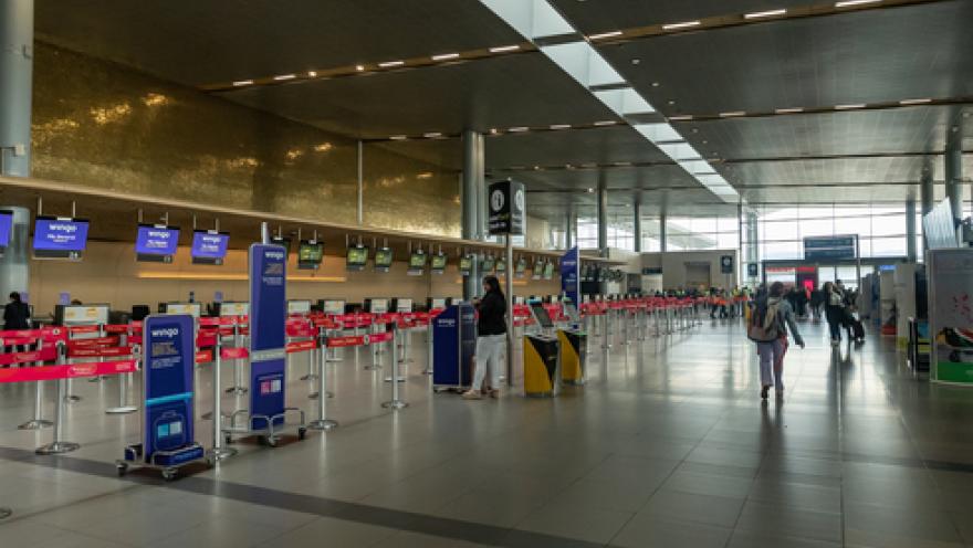 Interior of an airport showing a deep hall with several customer service areas. Some people with suitcases are walking along the front.