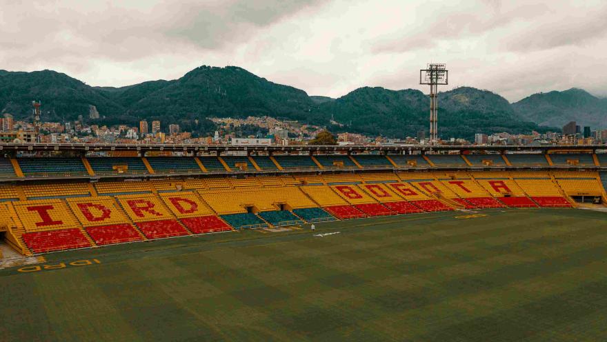 Interior del Estadio, desde arriba, se observa una tribuna de fondo pintada con las palabras IDRD y  Bogotá. Al fondo se alcanzan a ver los cerros orientales. 