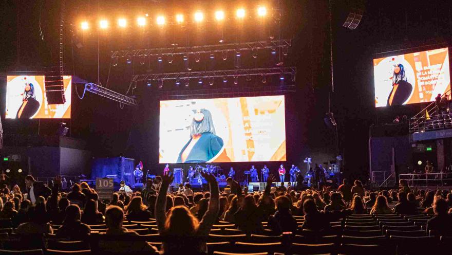 Escenario a oscuras con público sentado y un grupo artístico en la tarima. Se ven tres pantallas gigantes emitiendo un video.