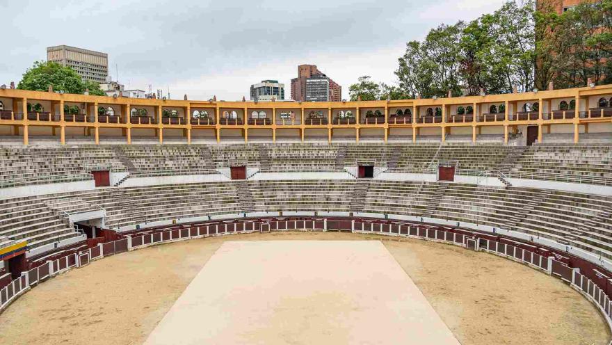 Interior de la plaza, se ve de frente y cerca la arena con una tabla en el centro, al fondo una gradería blanca ovalada de dos pisos; y un tercer piso, con balcones de color amarillo. Por encima y al fondo resaltan la parte alta de algunos edificios y unos árboles altos.