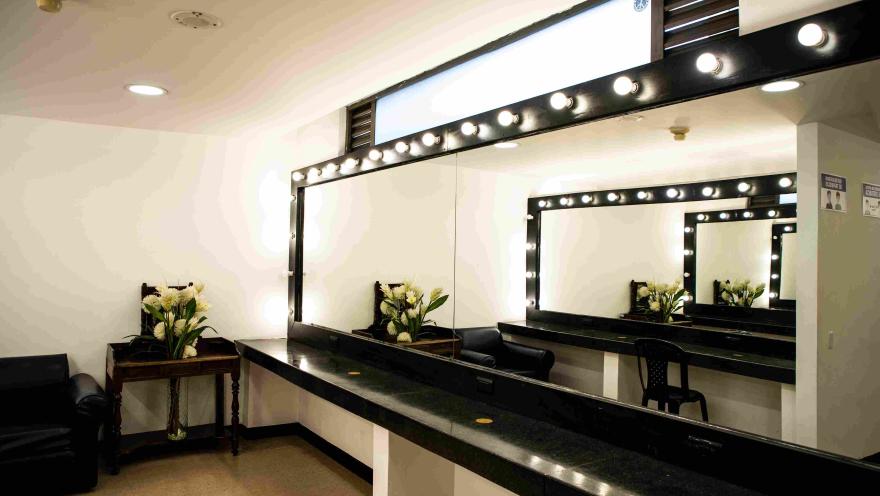 Interior del teatro en el que parece un camerino con una mesa de mármol larga y al frente de una pared en la que está un espejo rectangular del que sobresalen bombillas encendidas. Hay una flores en una mesa en una esquina.