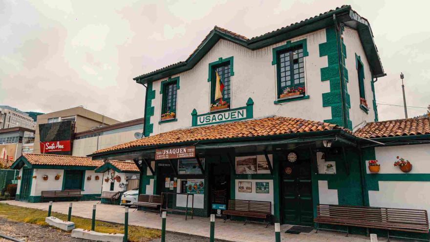 Estación de tren en Usaquén, con una casa que es la taquilla, con fachada blanca con rayas verdes, techo en teja de barro y dos avisos que dicen: "Usaquén".