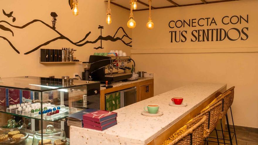 Interior de un local comercial cuyas paredes tienen color es castaño claro y en donde hay una vitrina con chocolates y tortas, y una grán máquina para preparar café. 