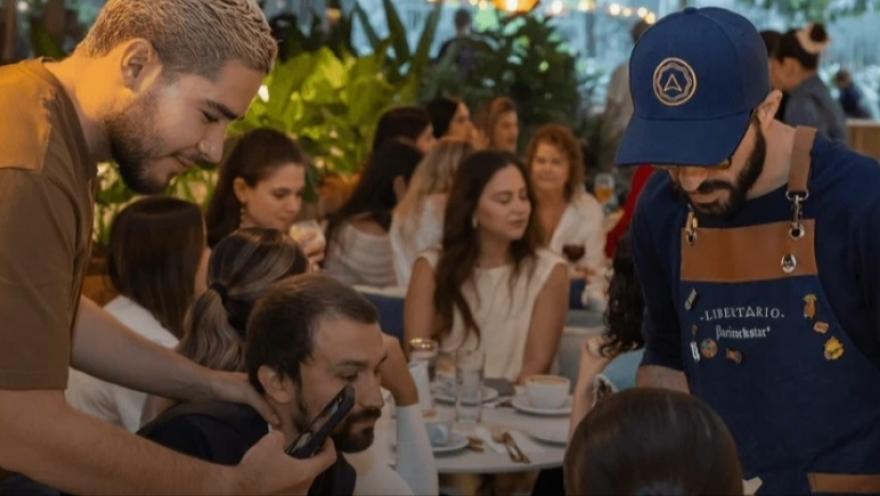 A group of young people in a restaurant is listening to a young waiter wearing a blue apron.