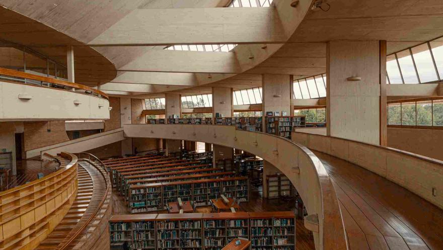 Interior de biblioteca en la que destacan corredores de madera en círculos, ventanales a los costados y abajo un salón con varias estanterías repletas de libros.