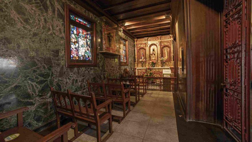 Interior de capilla con paredes y techos de madera y mármol. Hay una ventana en vitral cinco sillas de madera amplias y vacías. Al fondo hay un altar.