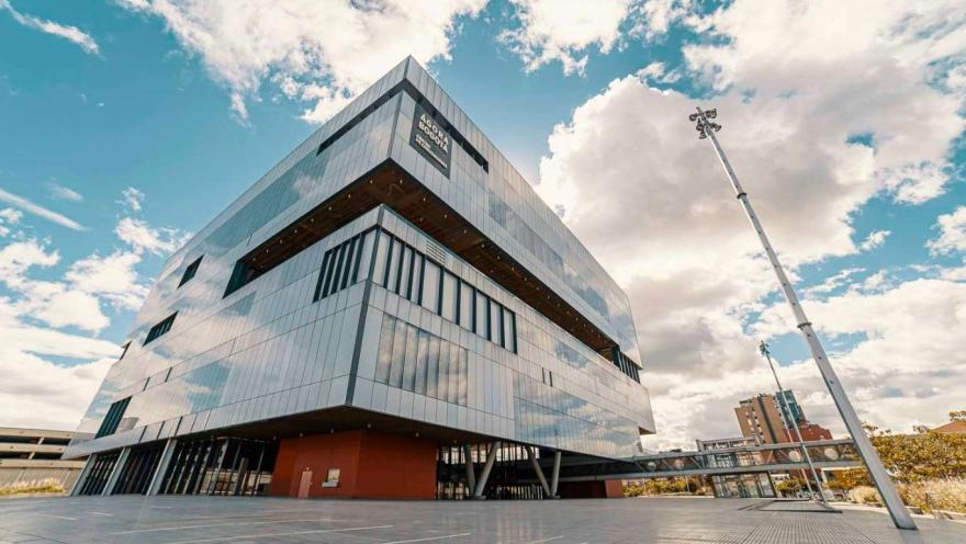  Parte exterior de un edificio cuyas paredes son grandes vidrios, al frente hay una plazoleta vacía. En la parte alta de la pared hay un aviso que identifica al edificio en el que se lee "Ágora Bogotá"
