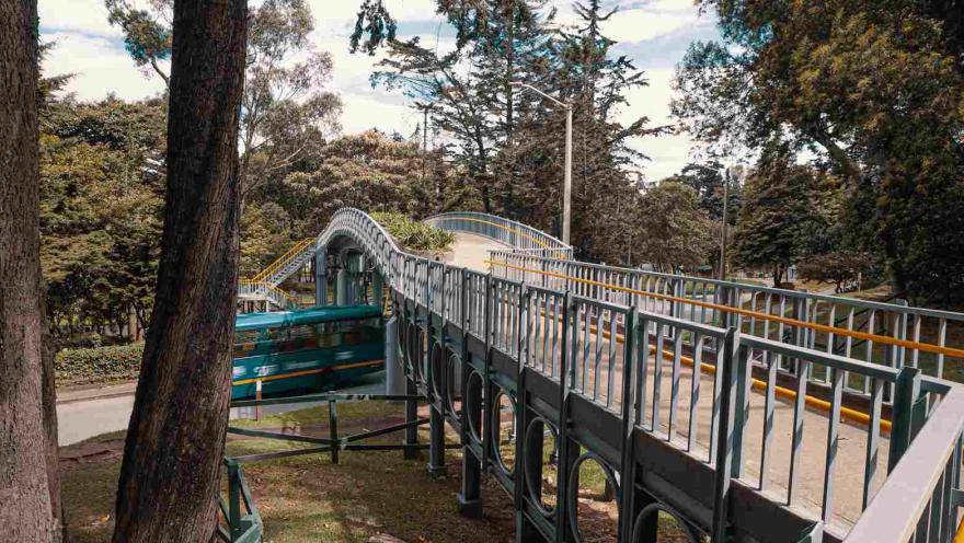  Puente de hierro con figuras geométricas sobre una avenida por la que pasa rápido un bus. Está rodeado de altos árboles.