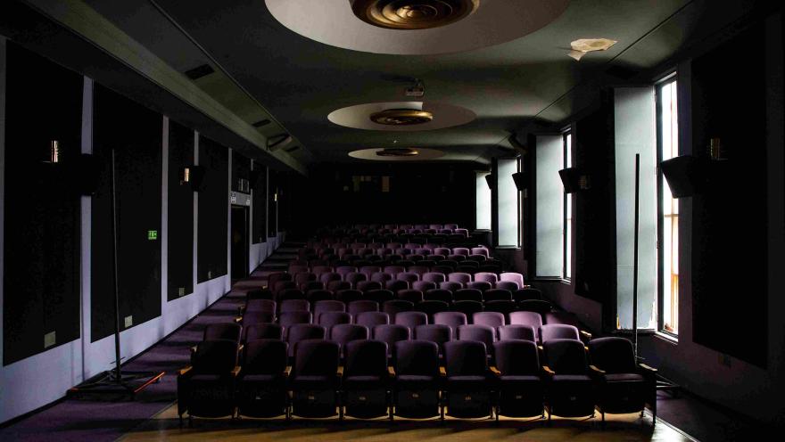 Salón como de cine con varias filas de sillas color violeta, de frente a fondo, lámparas inclrustadas en el techo y a un lado 4 ventanas abiertas con luz de día afuera.