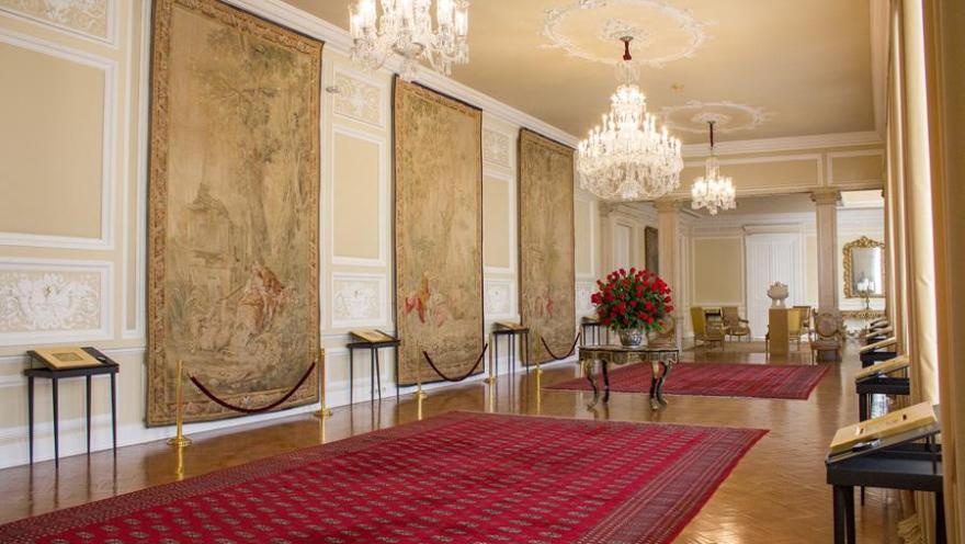 Salón largo con tapetes rojos y 3 tapices en una pared. Hay tres lámparas de cristal en el techo.