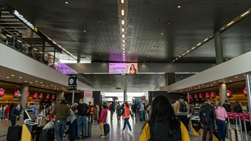 Interior of an airport showing a deep hall and some people with suitcases walking along the front.