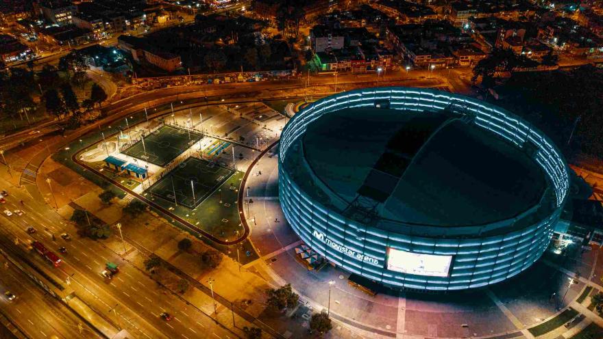 Panorámica nocturna vista desde arriba del Movistar Arena de noche. Se ve un par de chachas sintéticas, y al fondo unas edificaciones de barrio iluminadas. 
