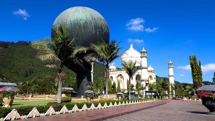 Monumento la Mano de Dios, una mano que sostiene una esfera que parece ser el mundo, y al fondo la réplica del Taj Mahal. Varias palmeras y  una montaña de fondo adornan el entorno de un soleado día.