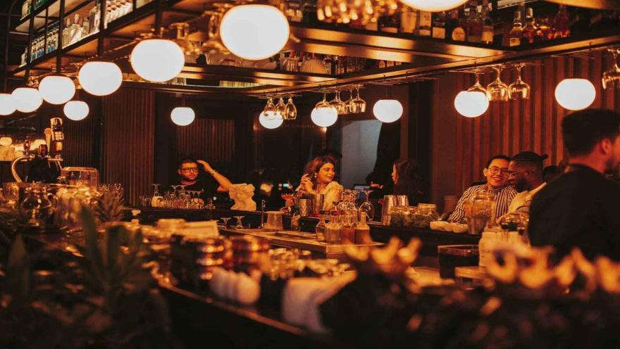  Un bar animado y concurrido, iluminado cálidamente, donde la gente disfruta de bebidas y conversaciones.