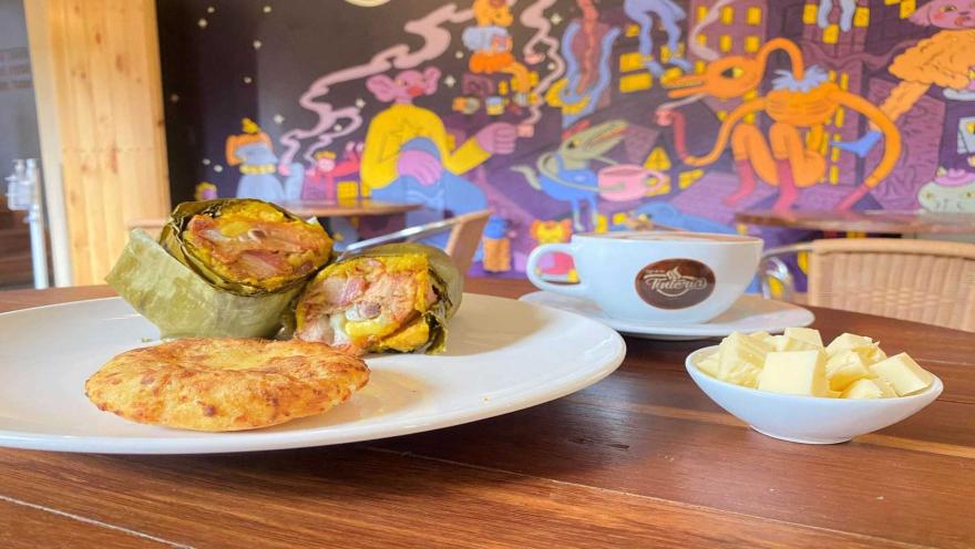Un tamal envuelto en hoja, una arepa de maíz, una taza de café y un tazón de queso, en una mesa de madera.