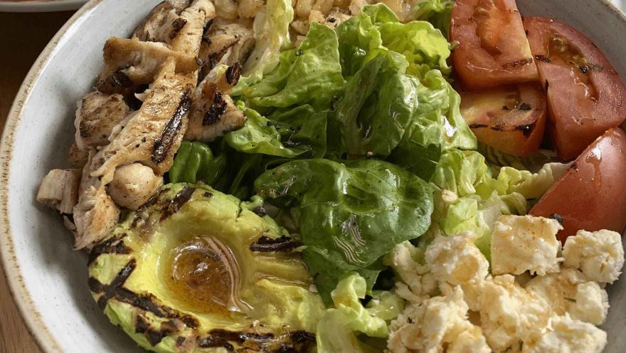 Ensalada fresca con pollo a la parrilla, aguacate, tomate, lechuga y queso feta, aderezada con vinagreta.