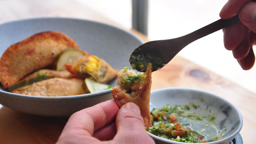 Manos sirviendo salsa chimichurri sobre una empanada frita, con un plato de empanadas al fondo.