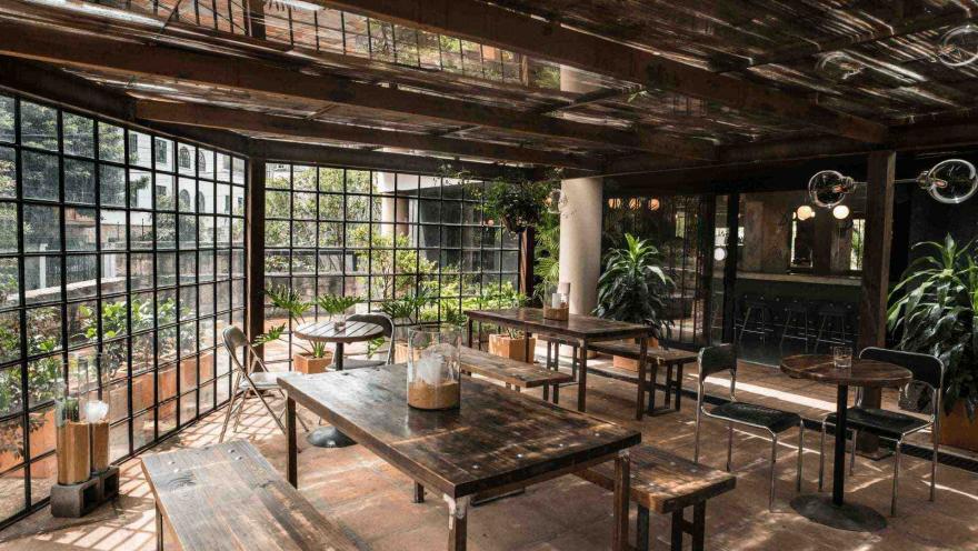Un comedor de restaurante con techo de cristal, mesas y sillas de madera, y plantas verdes.