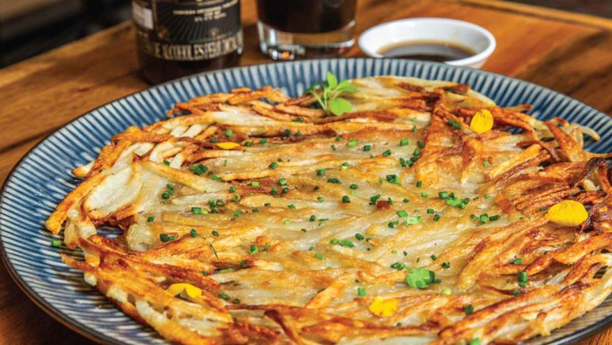 Una tortilla de papas ralladas y crujientes, espolvoreada con cebollín, presentada en un plato de diseño con borde azul.